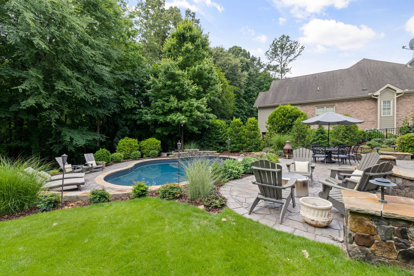 Jardin privé : piscine, terrasse en dalles de pierre avec fauteuils et parasol, massifs de graminées et pelouse sous de grands arbres